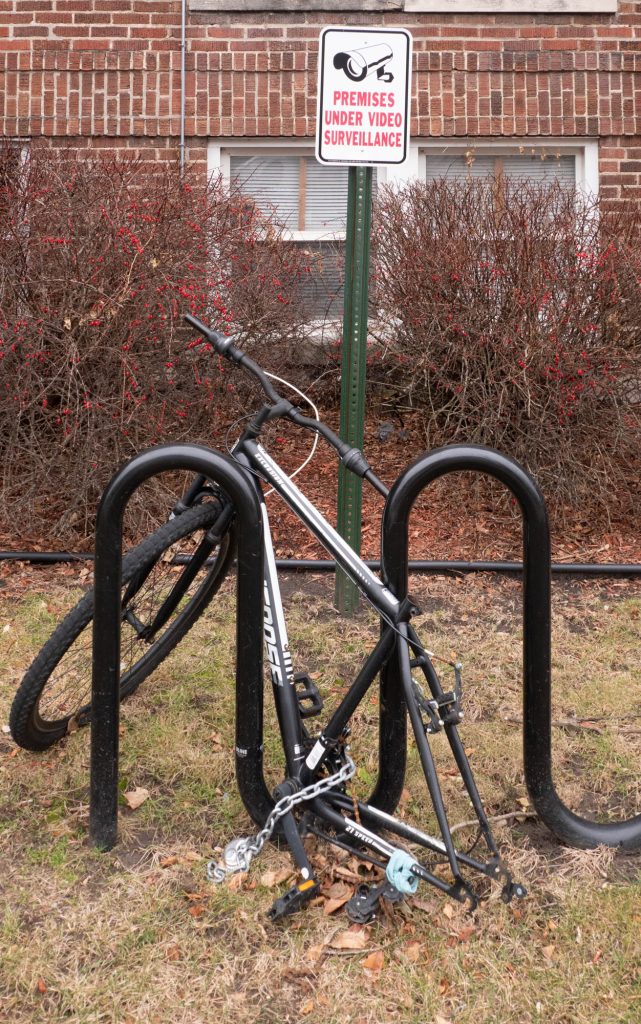Bike in rack with missing back wheel and seat.  Sign in background: Premises Under Video Surveillance