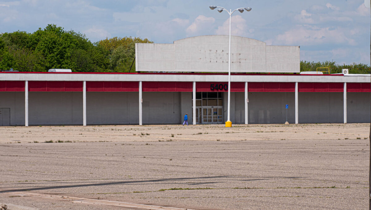 A figure in a blue coat walks in from of an abandoned K-Mart.